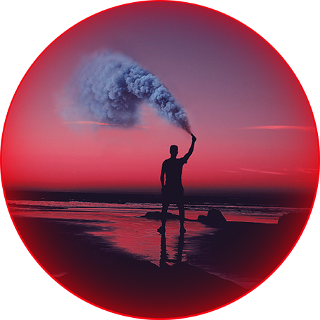 A man stands on the beachfront with his right hand raised, holding a flare that emits blue smoke.
